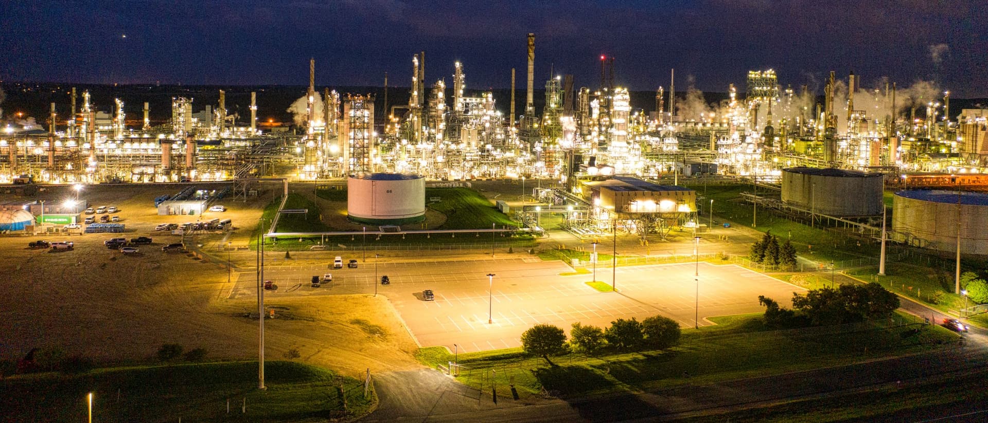 Vista aérea noturna de uma refinaria iluminada, com tanques e unidades de processo
